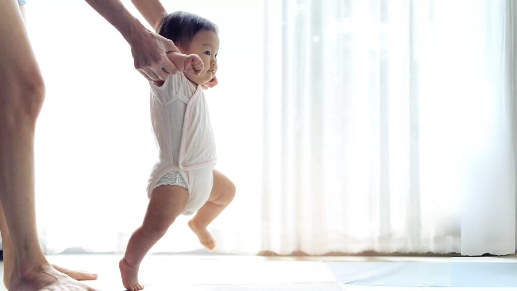 A baby taking assisted steps while holding onto an adult's hands, focusing on early motor skill development. This image represents paediatric physiotherapy techniques that help infants build strength, balance, and coordination for walking.
