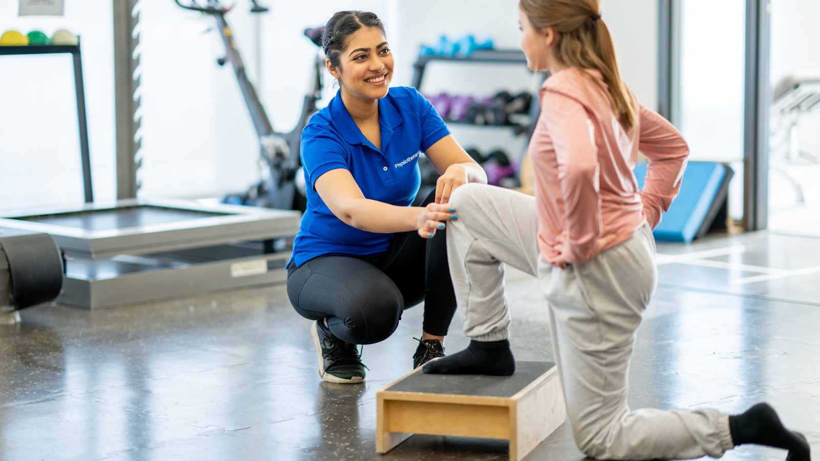 A female physiotherapist assisting a patient through a step-up rehabilitation exercise in a physiotherapy clinic setting.