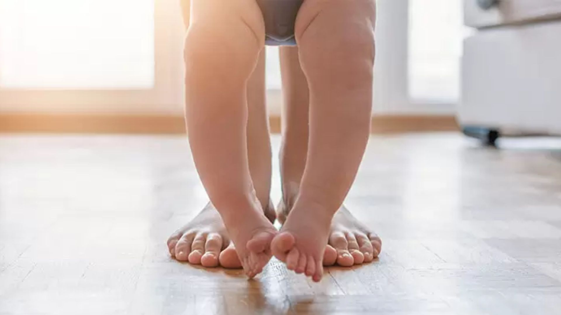 Baby standing on a parent’s feet in natural sunlight, learning balance and early standing.