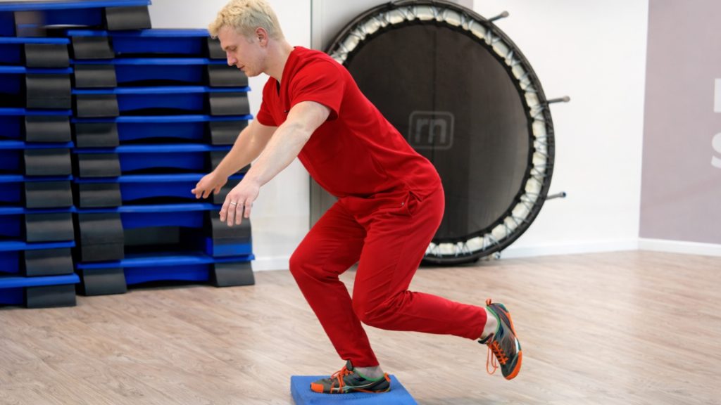 Physiotherapist demonstrating a single-leg balance and knee strengthening exercise in a clinic.
