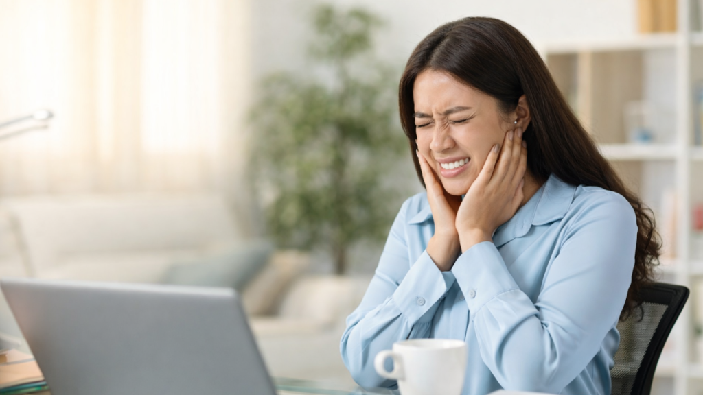 Asian office worker holding her jaw in pain while working on a laptop, illustrating stress-related jaw pain.