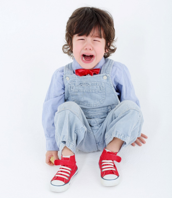 A young boy wearing overalls and red shoes is crying