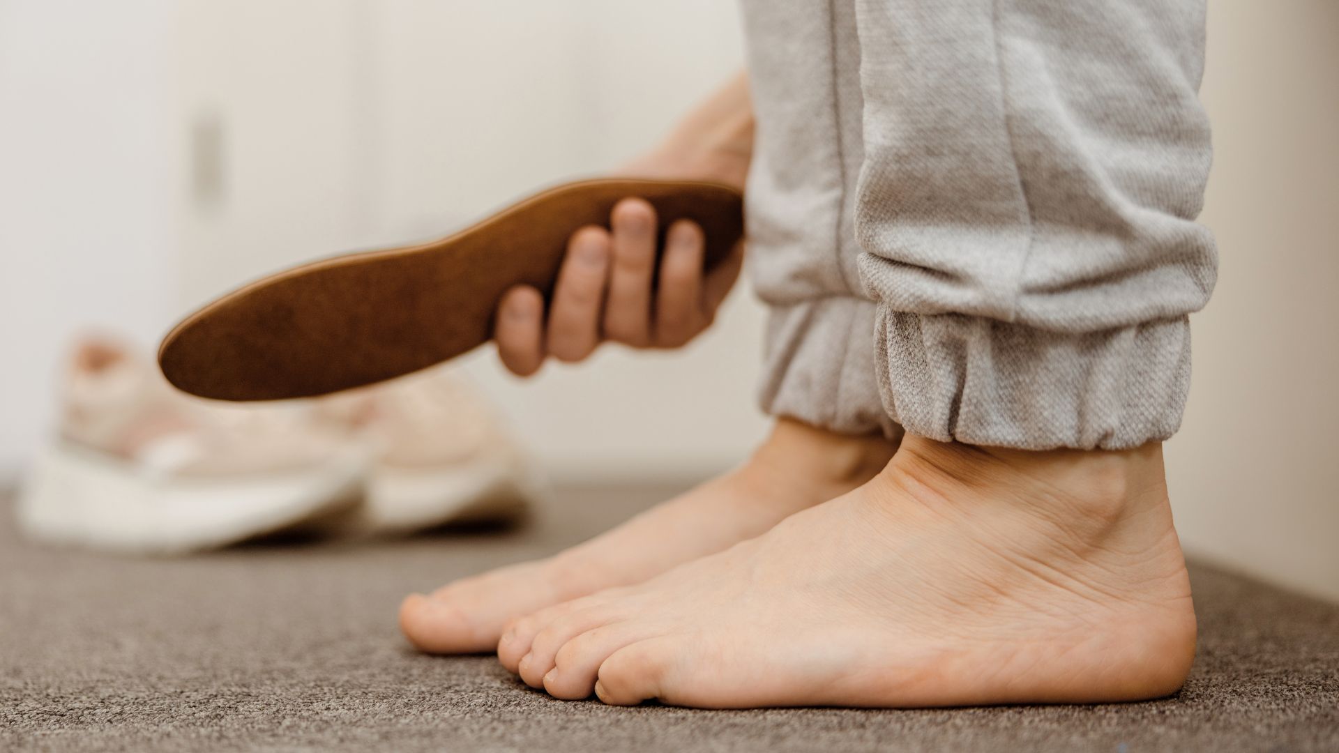 Person holding a custom insole while standing barefoot on the floor, highlighting foot support and orthotic solutions for better comfort and alignment.