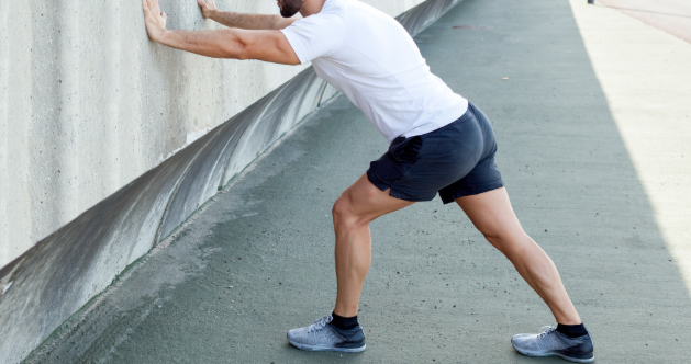 Man performing a calf stretch against a wall, a common exercise to improve flexibility and relieve tightness in the lower leg muscles. This stretch is often recommended for runners and individuals with foot and ankle conditions such as plantar fasciitis or Achilles tendonitis.