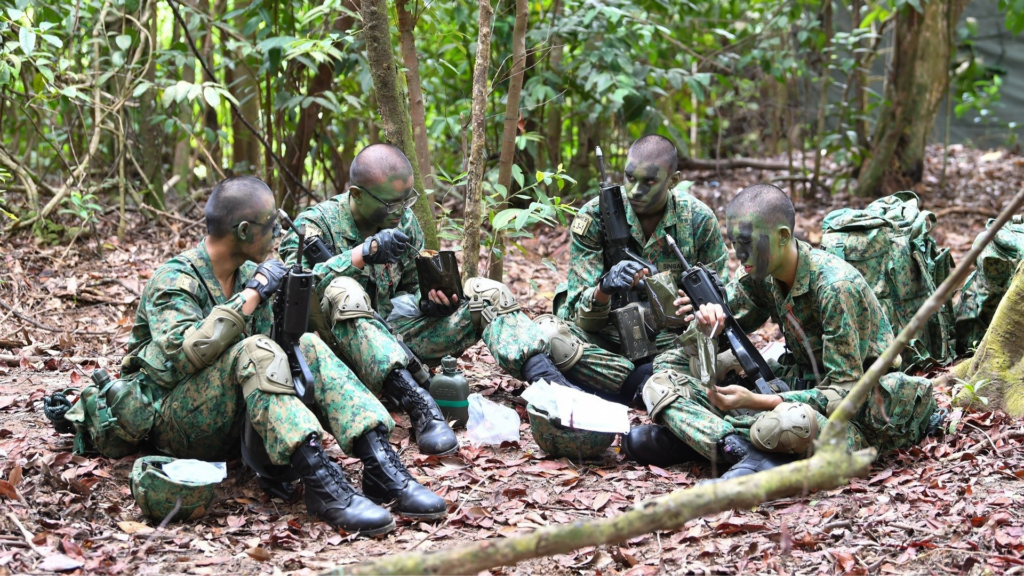 SAF recruits in camouflage gear during field camp, sitting on forest ground; highlights importance of physio support for injury prevention in tough terrains.