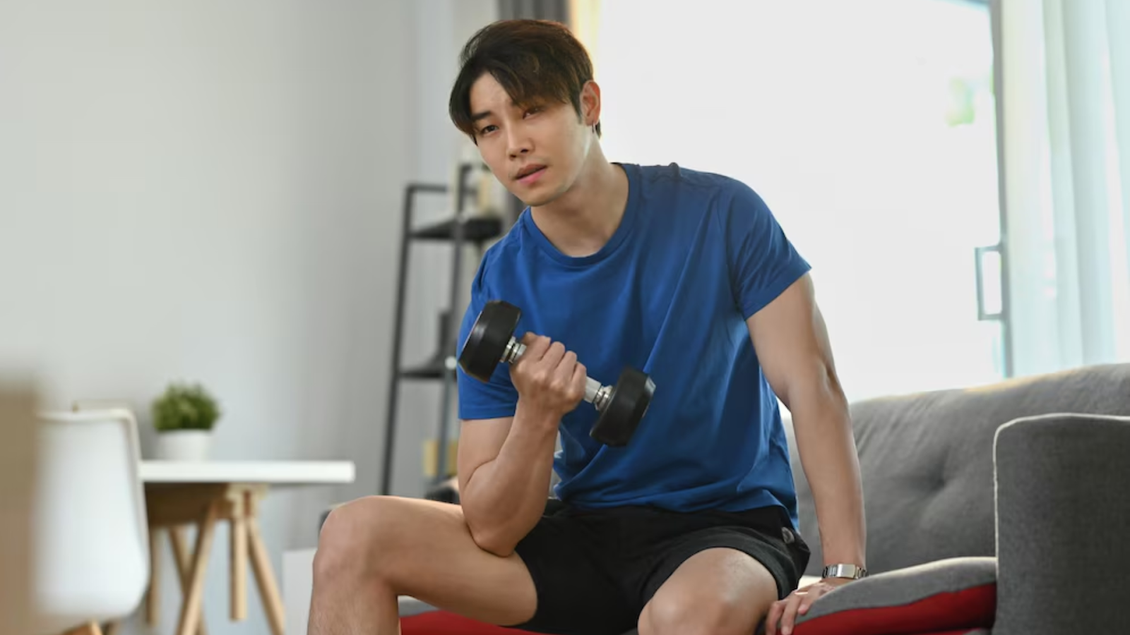 Young man sitting on a sofa doing bicep curls with a dumbbell at home