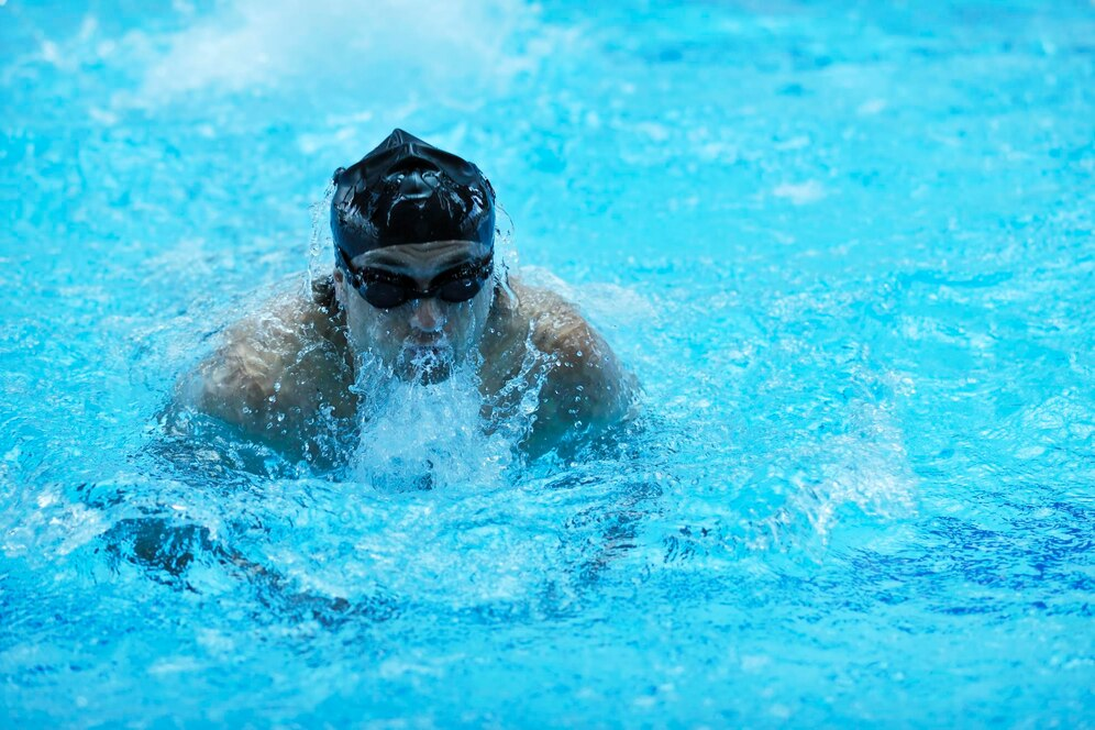 Competitive swimmer in black swim cap and goggles performing breaststroke in a bright blue pool.