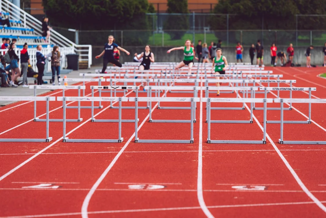 Female athletes sprinting and jumping over hurdles during a track and field race.