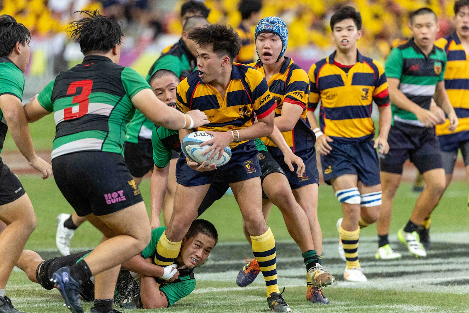 School rugby team in striped jerseys competing in a match with active tackles and crowd in the background.