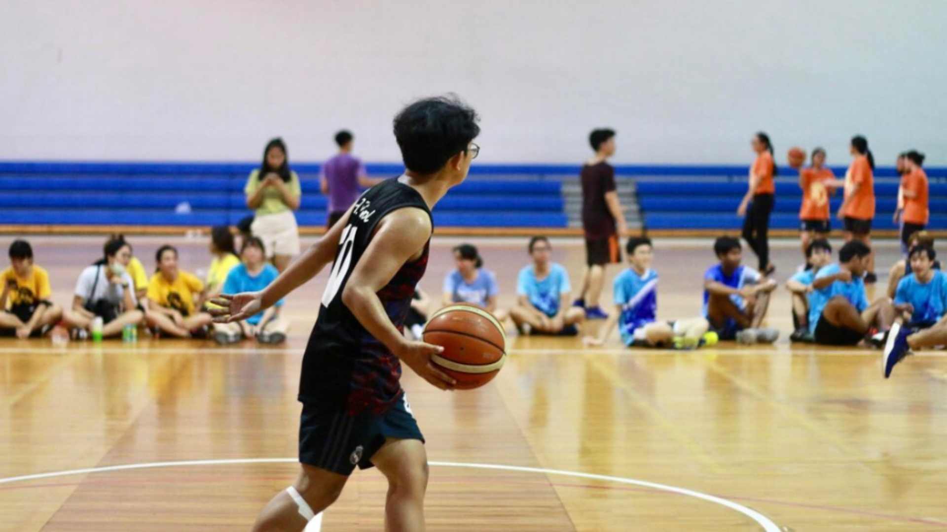 Teenage boy dribbling a basketball during a school match while spectators watch from the sidelines.