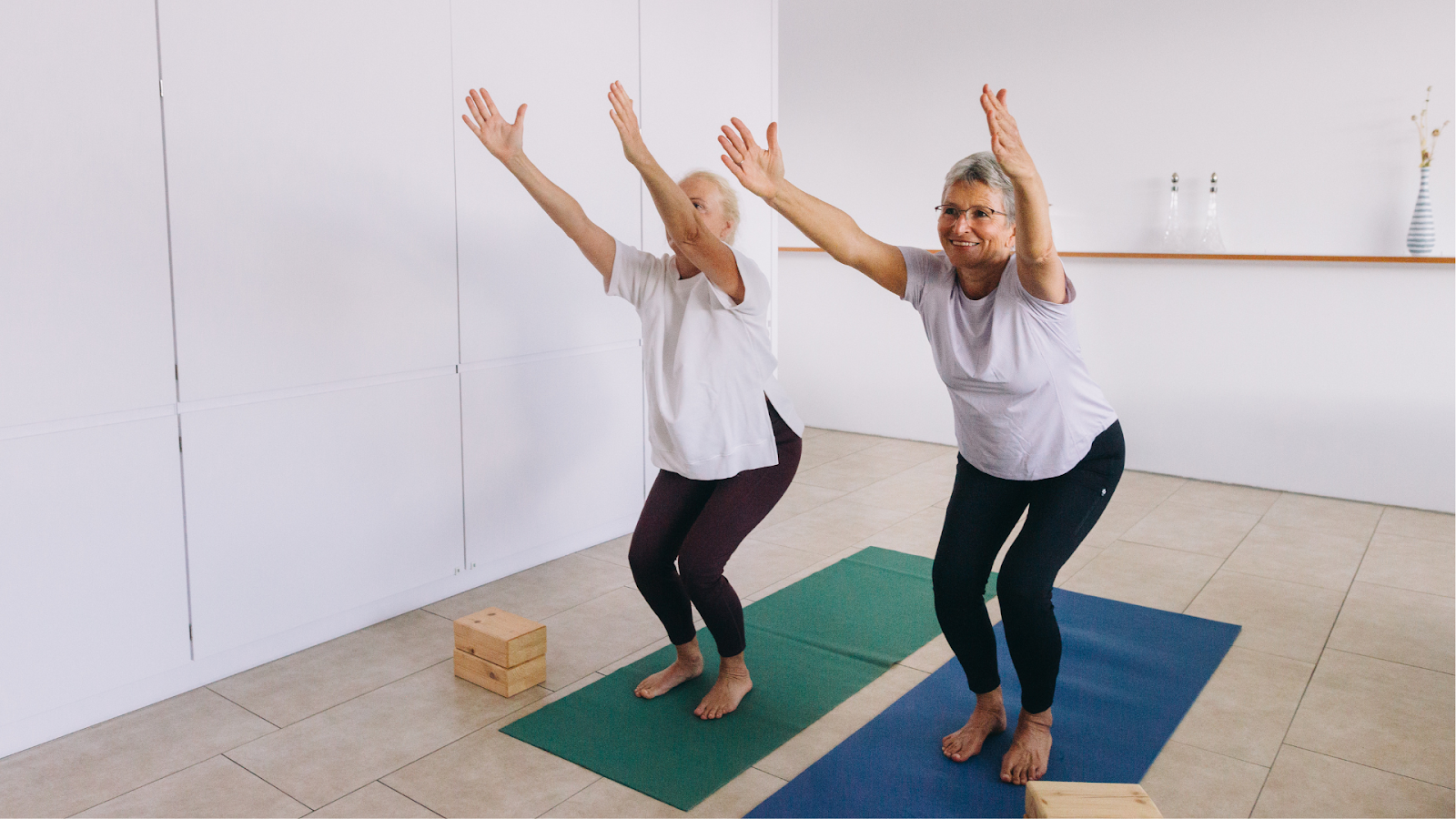 Two senior women doing a balance and strengthening physiotherapy exercise together indoors.