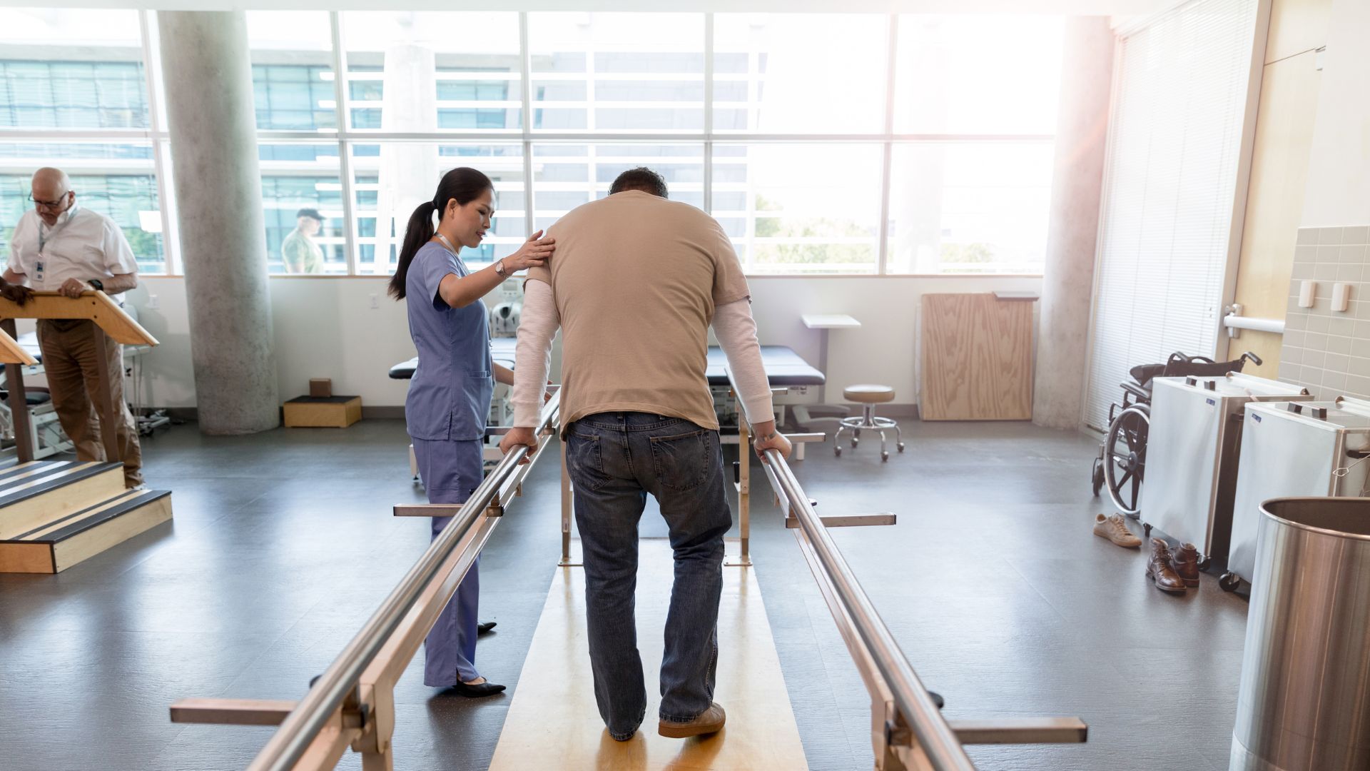 Physical therapist assisting a patient walking between parallel bars during rehab post-surgery.
