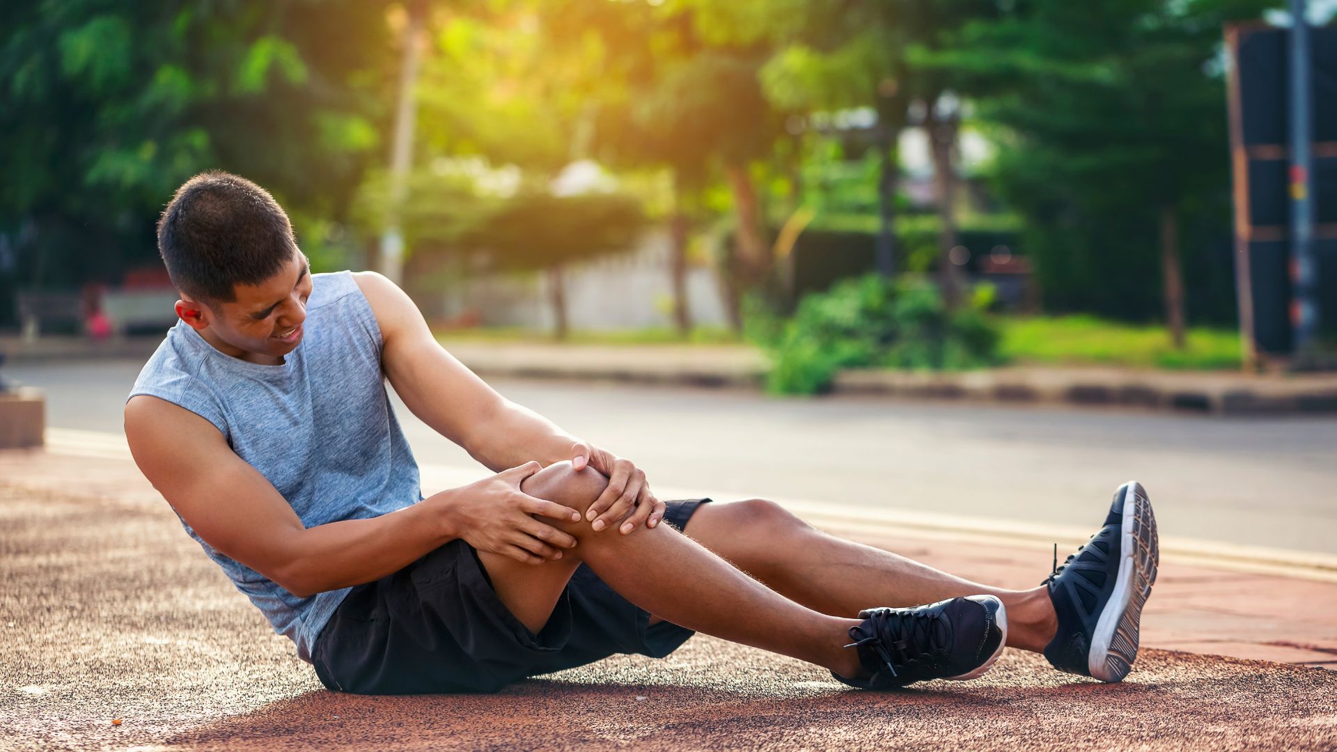Outdoor image of an active individual holding their knee in pain while seated on a track, representing common sports injuries addressed by sports physiotherapy.