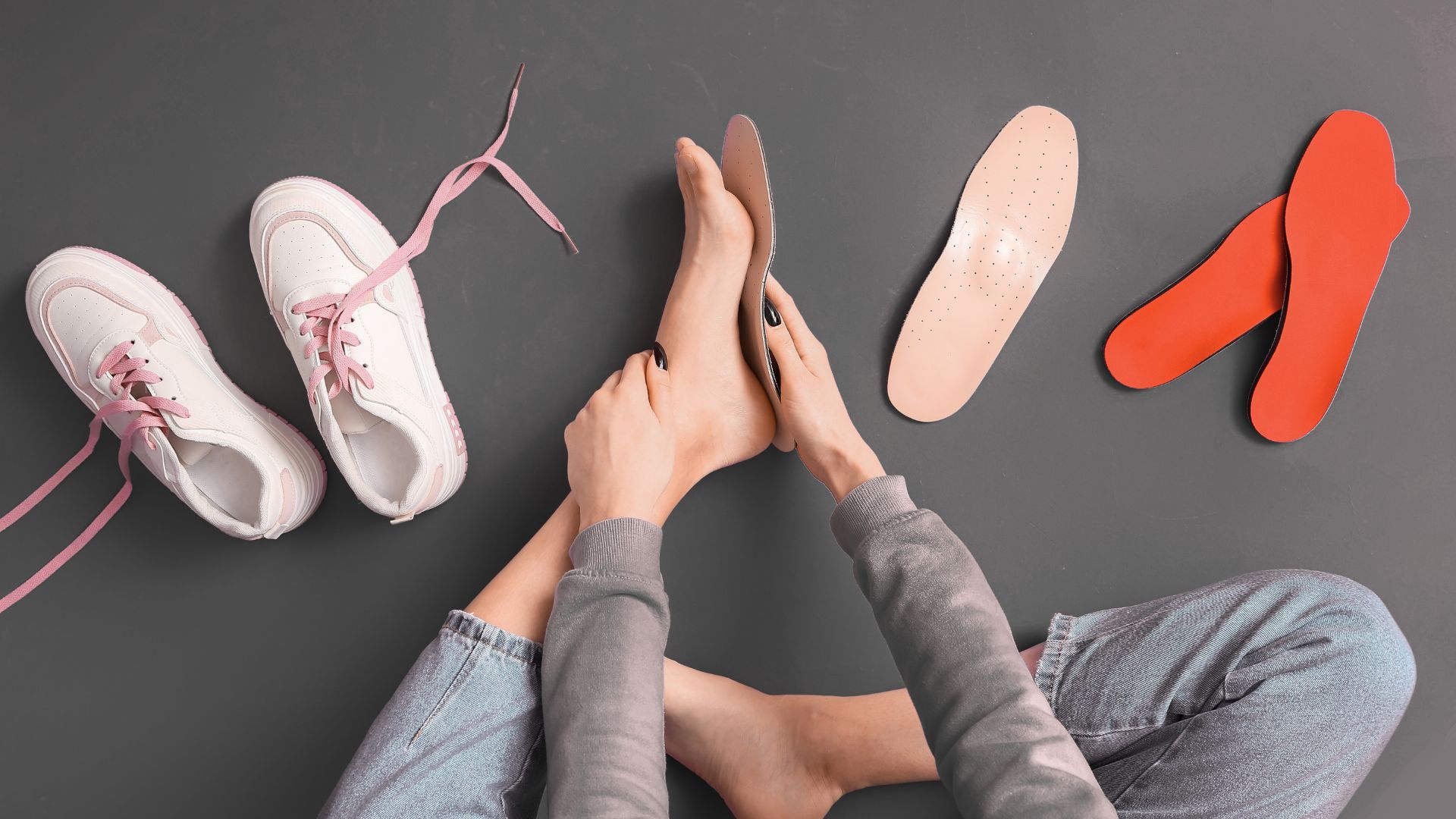 Person fitting shoe insoles onto bare feet, with sneakers and orthotics on the floor.