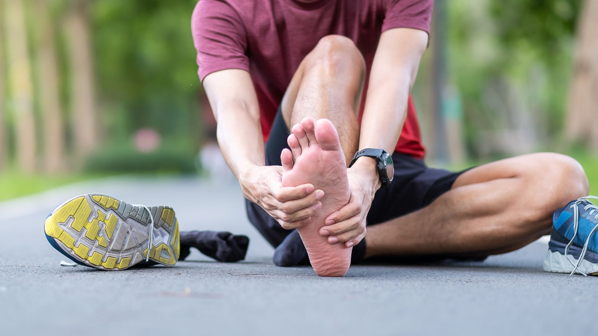 A man sitting on a park pathway holding his bare foot in pain, likely due to plantar fasciitis, with running shoes beside him.