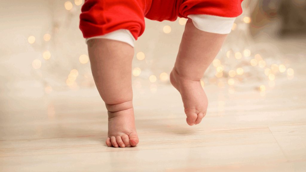 Close-up of baby’s chubby feet tiptoeing indoors on a wooden floor.
