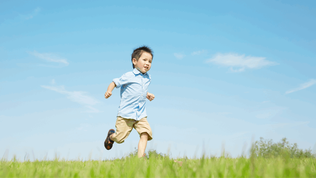Young boy running happily on a grassy field under blue sky.