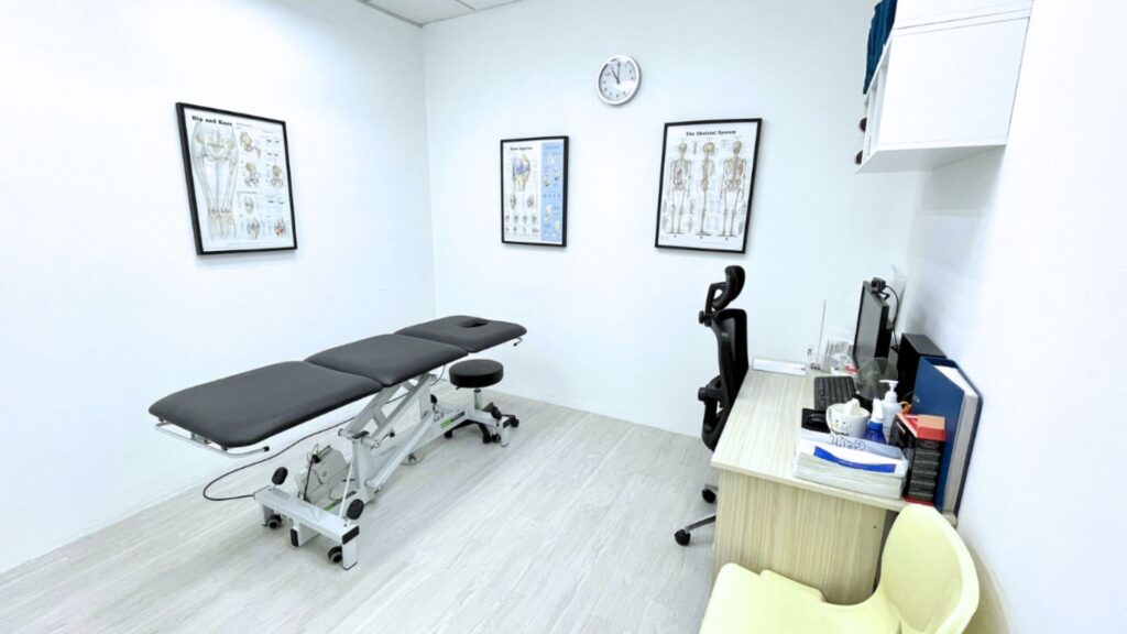 Clean and minimalist consultation room at Physio & Sole Clinic Kembangan featuring a treatment bed, anatomical charts, and a desk setup for patient assessments.