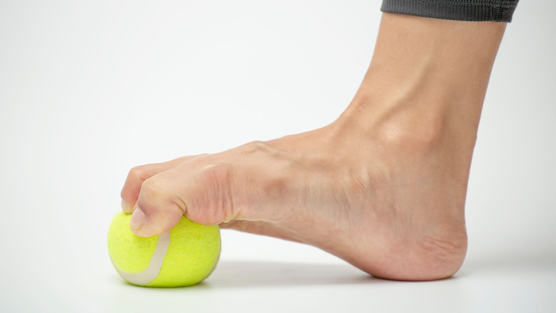 A person pressing their foot onto a tennis ball, showing a common exercise used to strengthen the arch and improve mobility in high-arched feet.