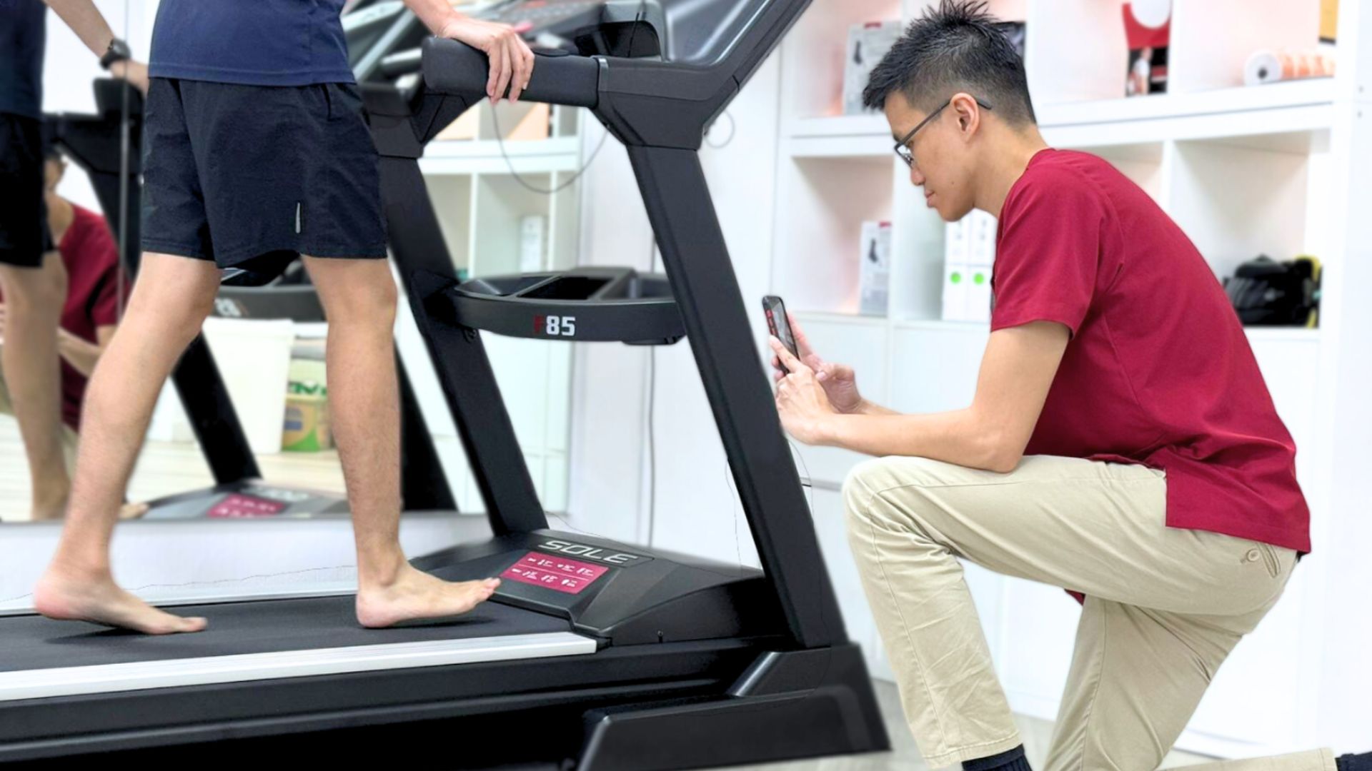 Podiatrist capturing gait analysis footage of a patient walking barefoot on a treadmill during a biomechanical assessment in Singapore.