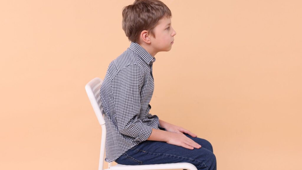 Child with rounded back sitting on a chair, illustrating posture issues that may stem from flat feet or poor lower limb alignment.