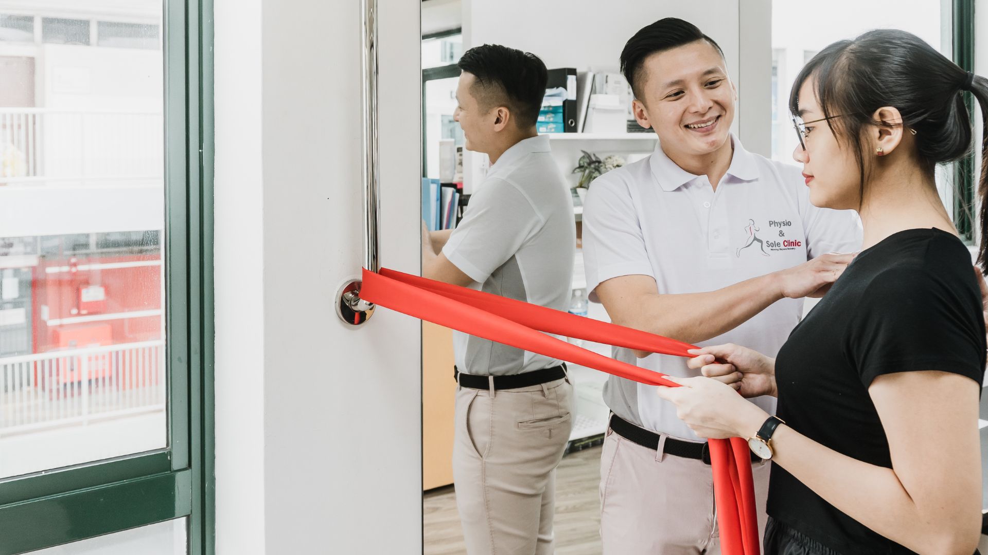 A physiotherapist guiding a patient through a shoulder strengthening exercise using a red resistance band in a Singapore clinic.