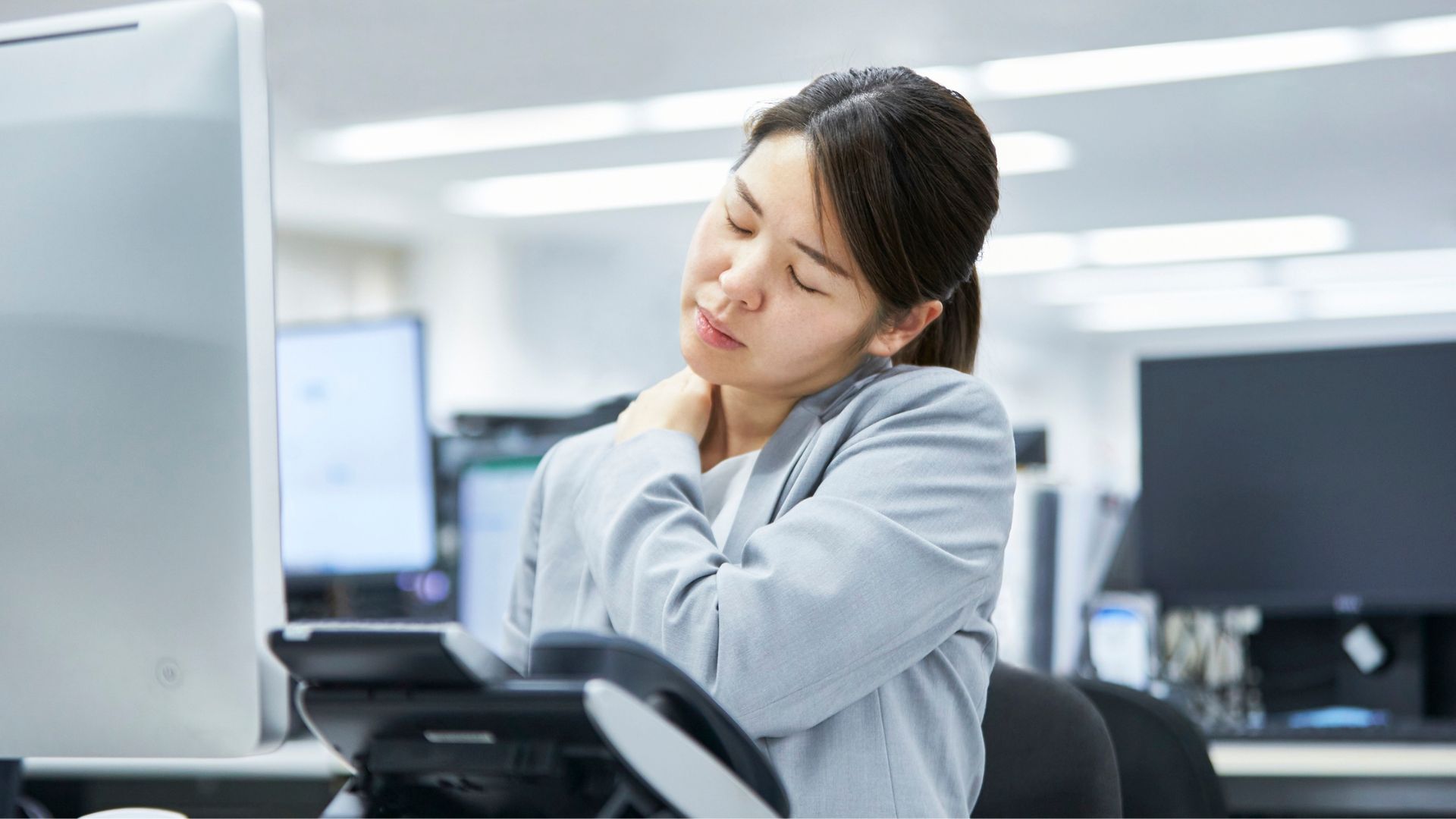 A woman experiencing neck and shoulder pain while working at her desk in an office setting.