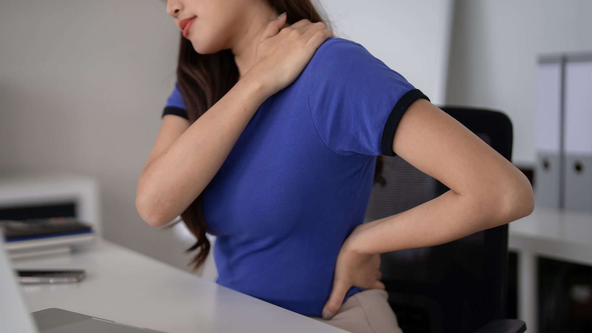 A woman holding her neck and lower back, showing discomfort from prolonged sitting at her workstation.