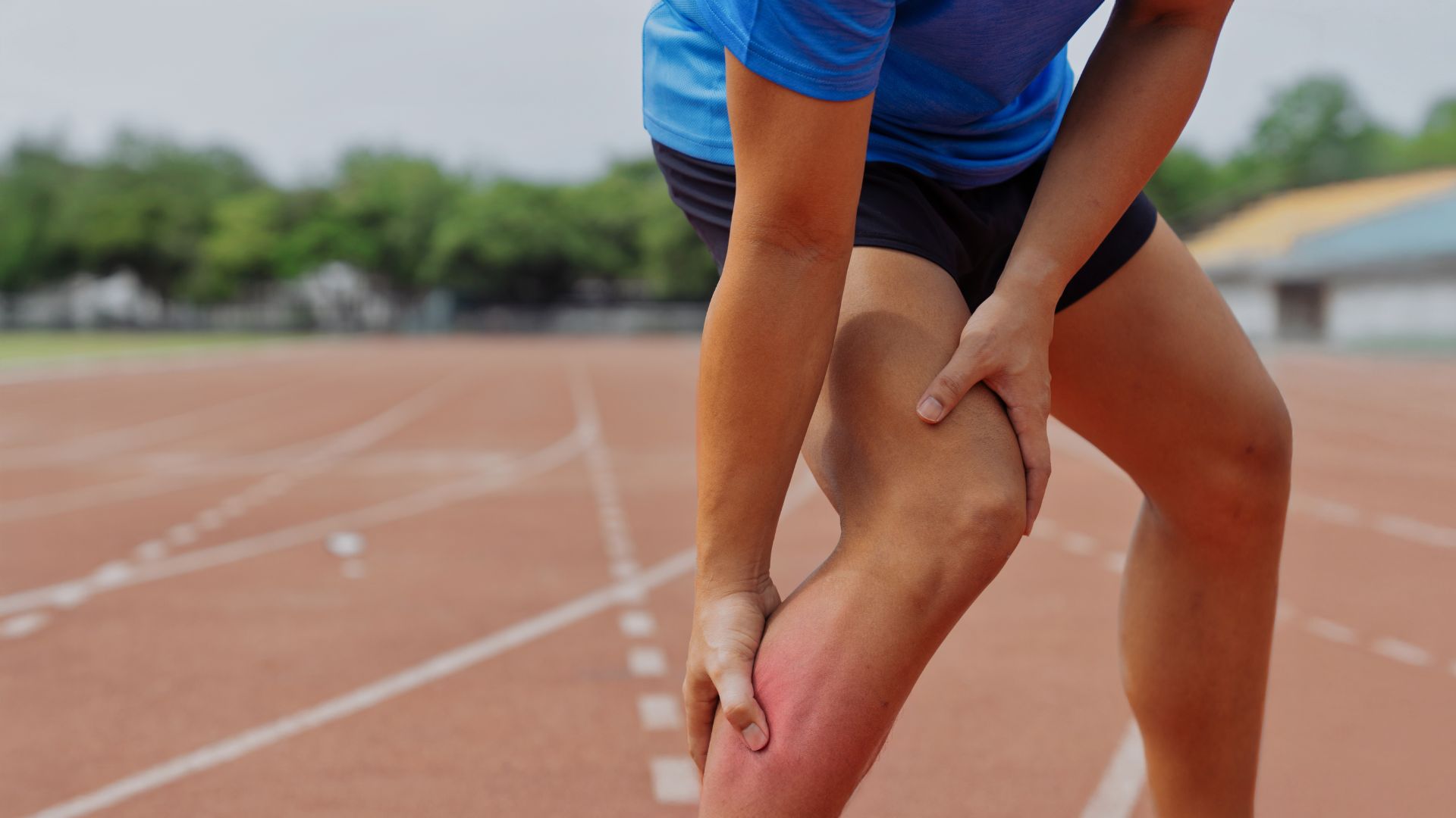A runner holding his thigh in pain on a running track, showing symptoms of a quadriceps strain or sports-related overuse injury.