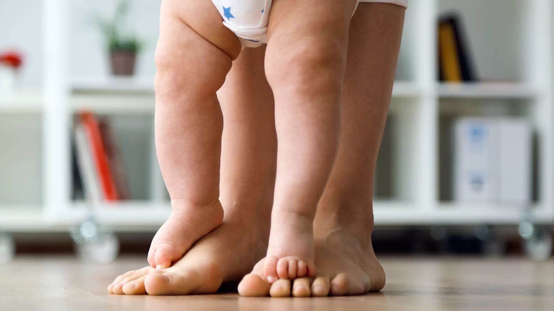 Baby standing on a parent’s feet indoors, demonstrating early standing posture.
