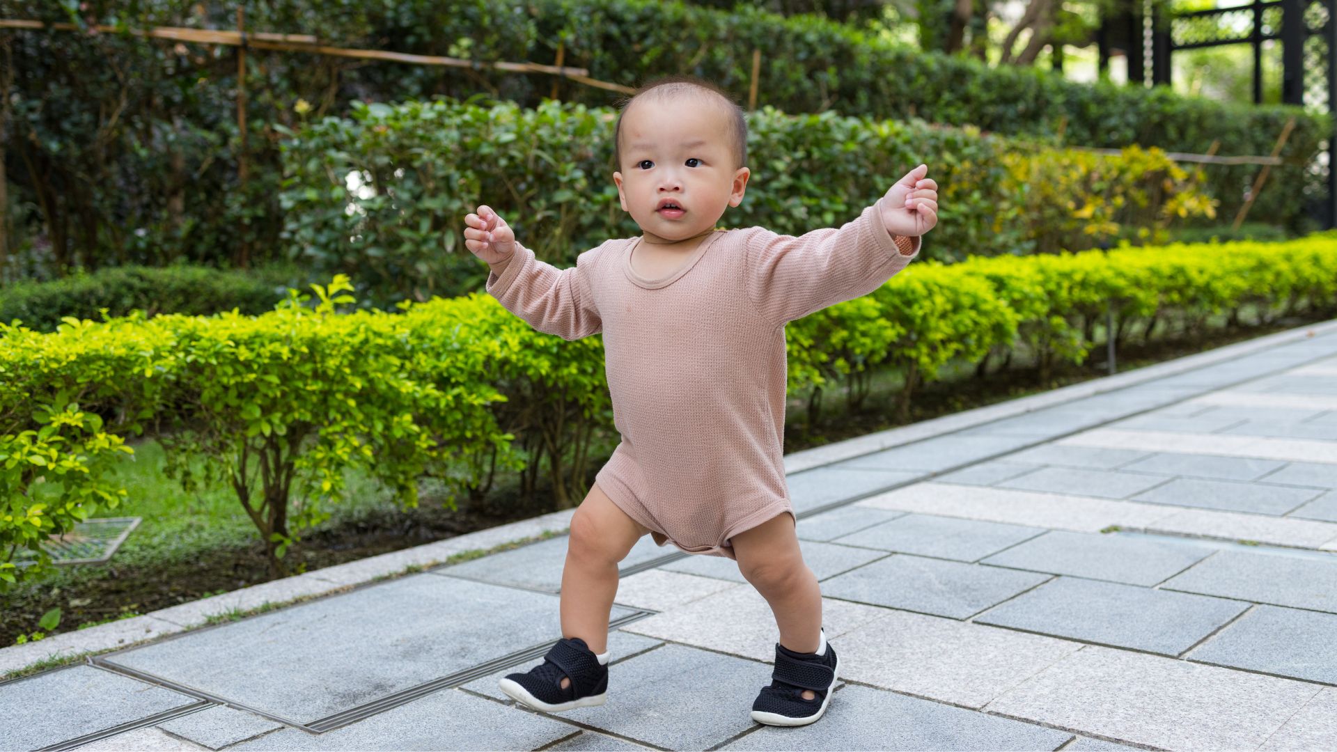 Toddler taking early steps outdoors with a wide stance, showing natural bow-legged posture.