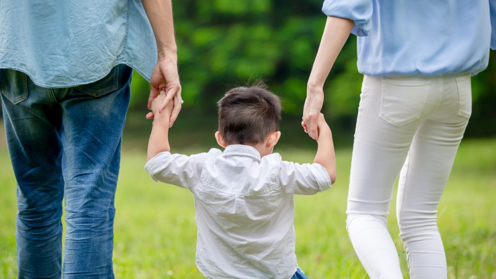 Parents holding their young child’s hands while walking outdoors, supporting early walking and gross motor development.