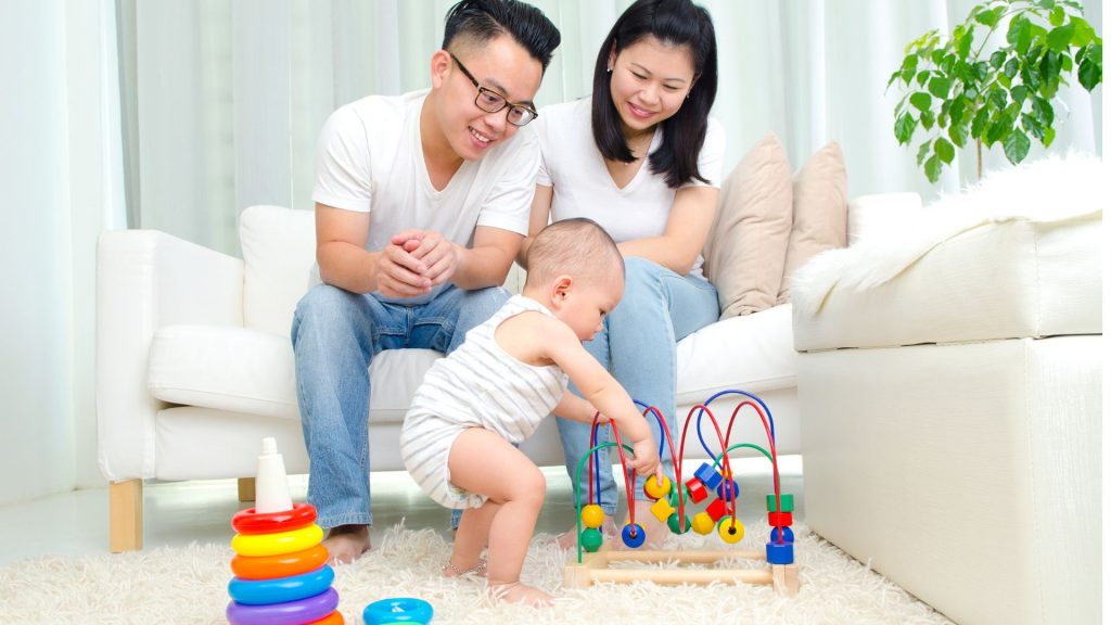 Parents in Singapore watching their baby stand and play with toys, supporting early motor development and balance.