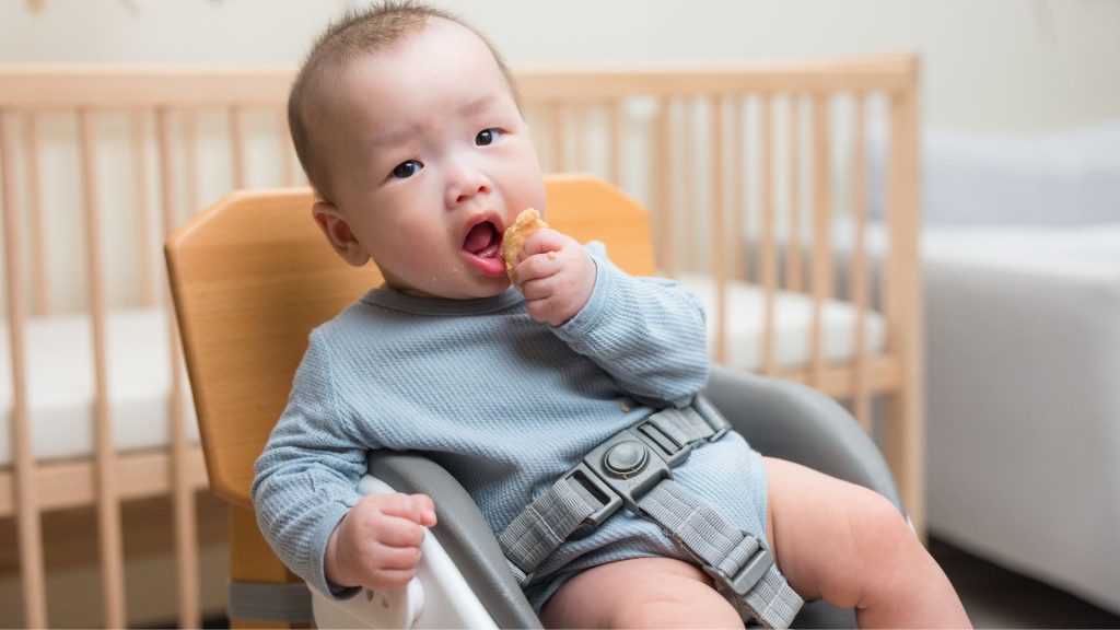 Baby sitting in a high chair feeding themselves, developing hand coordination and postural control.