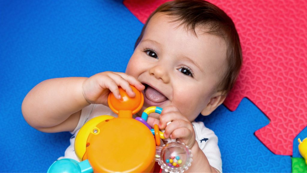 Baby lying on a play mat holding and mouthing a toy, developing fine motor skills and coordination.