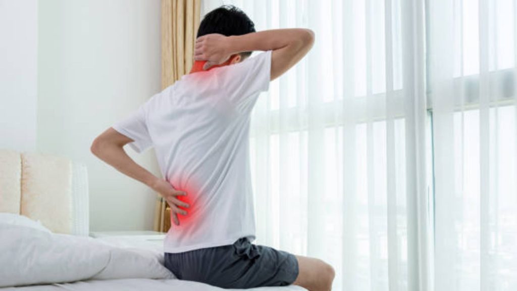 Man sitting on the edge of a bed holding his neck and lower back, with highlighted areas indicating neck and back pain commonly associated with muscle or posture-related discomfort.
