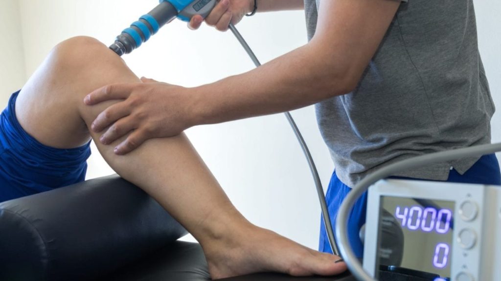Physiotherapist applying shockwave therapy to a patient’s knee using a handheld device, targeting tendon-related pain as part of evidence-based physiotherapy treatment.