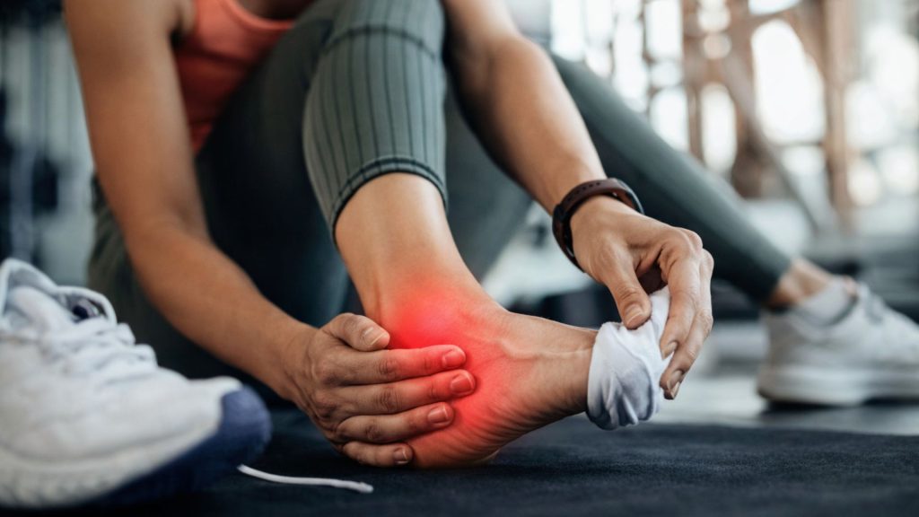 Gym user seated on the floor holding their heel with a red highlighted area, illustrating plantar fasciitis and heel pain after high-intensity or impact-based training.