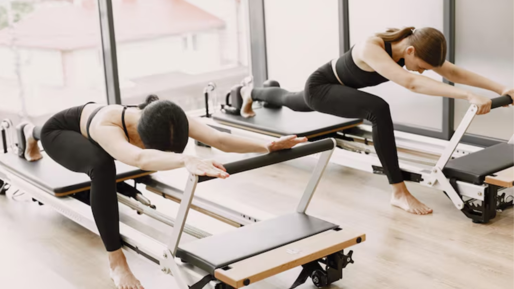 Two women performing Pilates reformer exercises in a bright studio, stretching forward with arms extended while kneeling and lunging on reformer machines to improve core strength, flexibility, and control.
