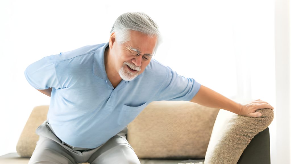 Senior man experiencing lower back pain while standing up from a sofa at home.
