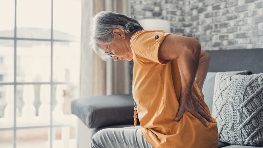 Elderly woman holding her lower back while experiencing back pain at home.
