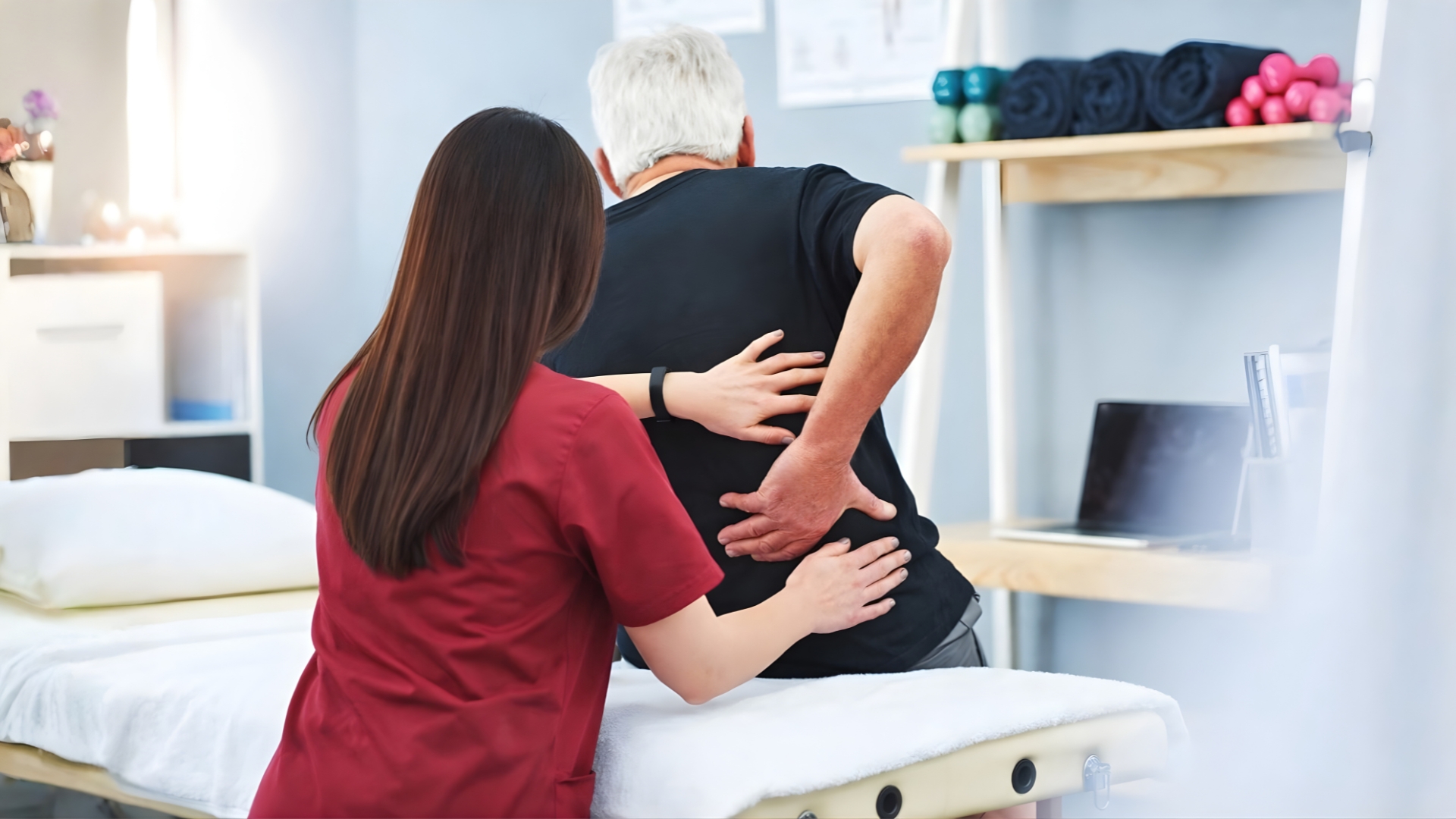 Physiotherapist supporting an elderly man’s lower back during musculoskeletal assessment in a clinic setting.