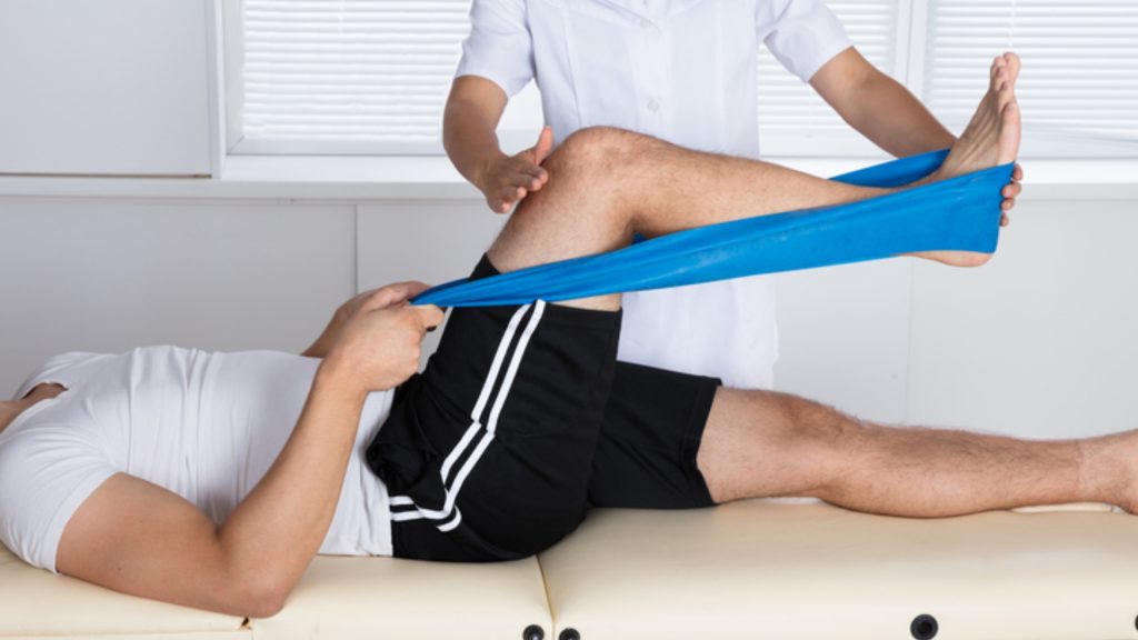 Therapist guiding patient through resistance band knee exercise on treatment table after ACL surgery.
