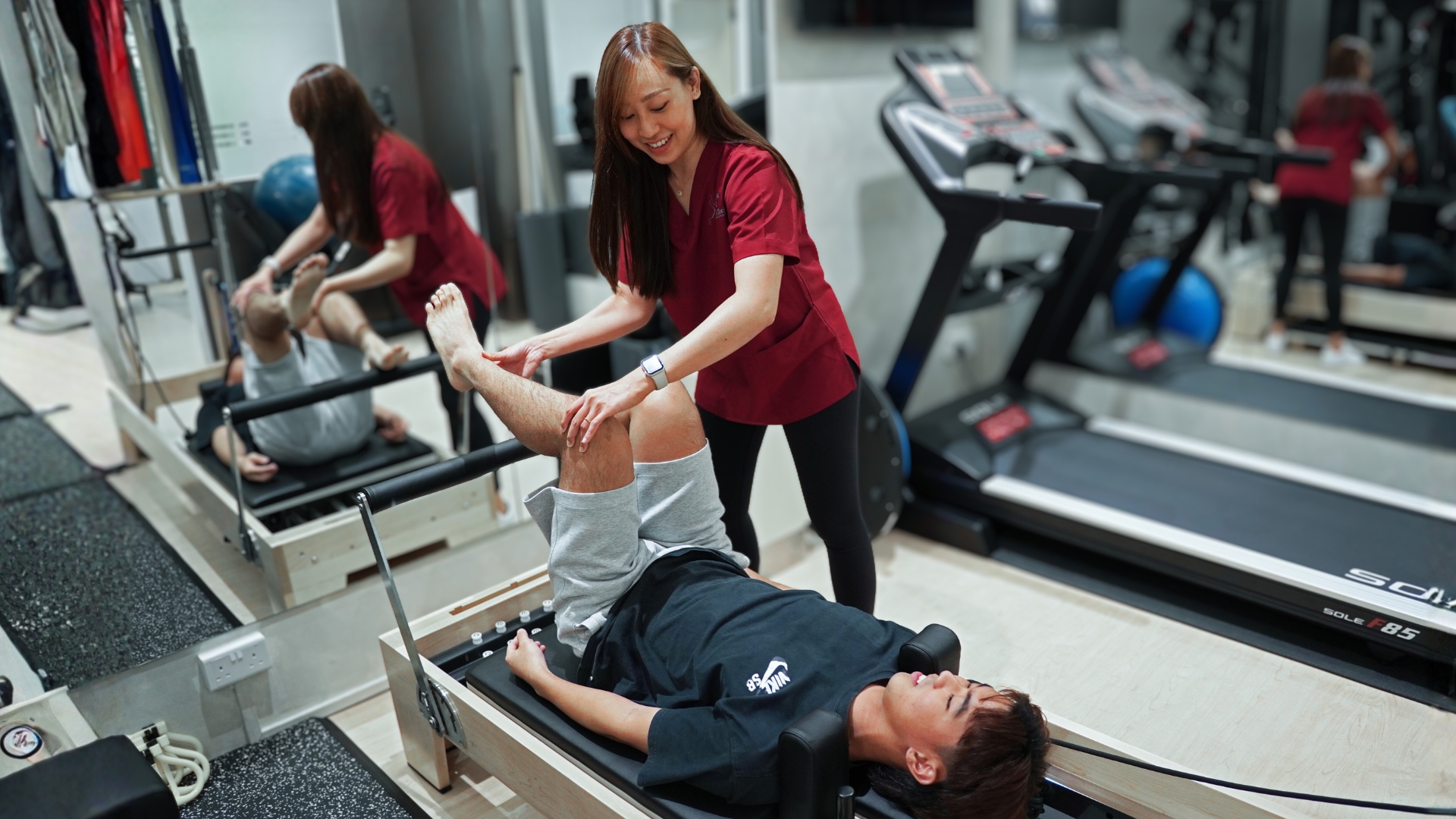 Physiotherapist assisting patient with leg strengthening exercise on reformer machine in rehab gym.