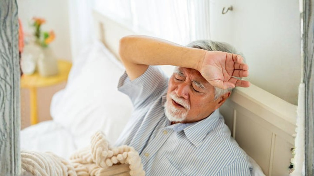 Elderly man sitting on bed looking fatigued and unwell at home.