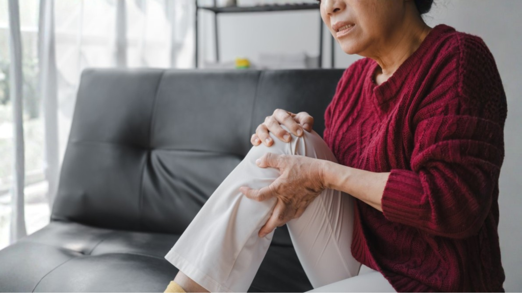 Senior woman sitting on sofa holding her knee in pain at home, indicating stiff joints.