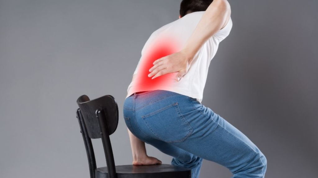 Person standing up from chair while holding lower back with highlighted area indicating lower back pain or sciatica