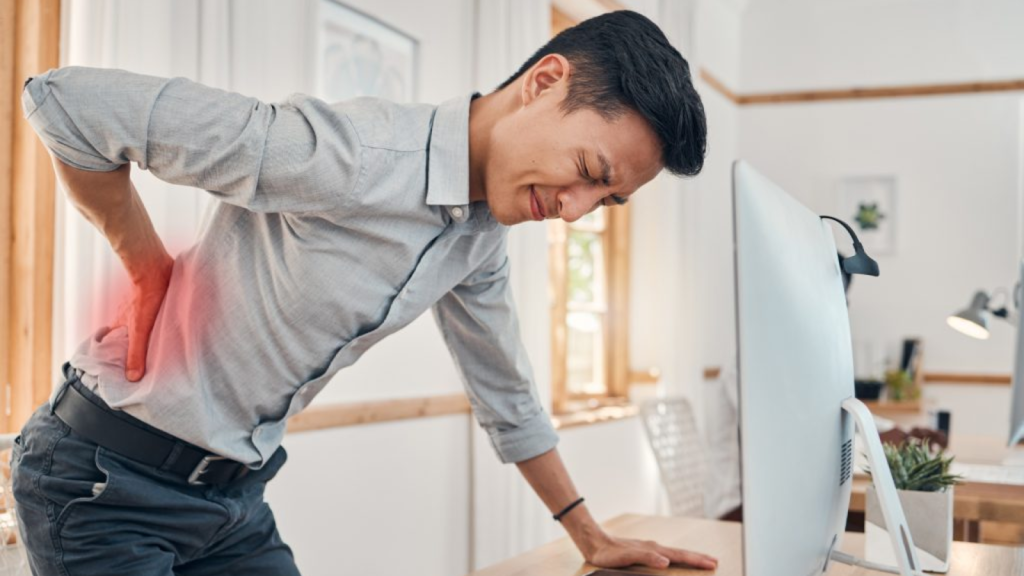Office worker holding lower back in pain while standing at desk with computer, showing lower back strain from prolonged sitting